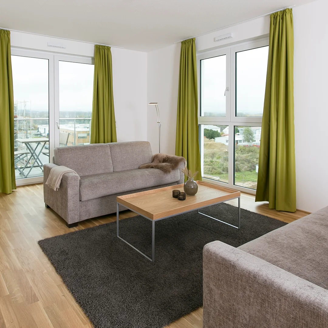 Appartement Budenheim Parkallee Wohnzimmer Wohnzimmer mit zwei grauen Sofas, einem Holztisch auf dunkelgrauem Teppich und grünen Vorhängen vor großen Fenstern.