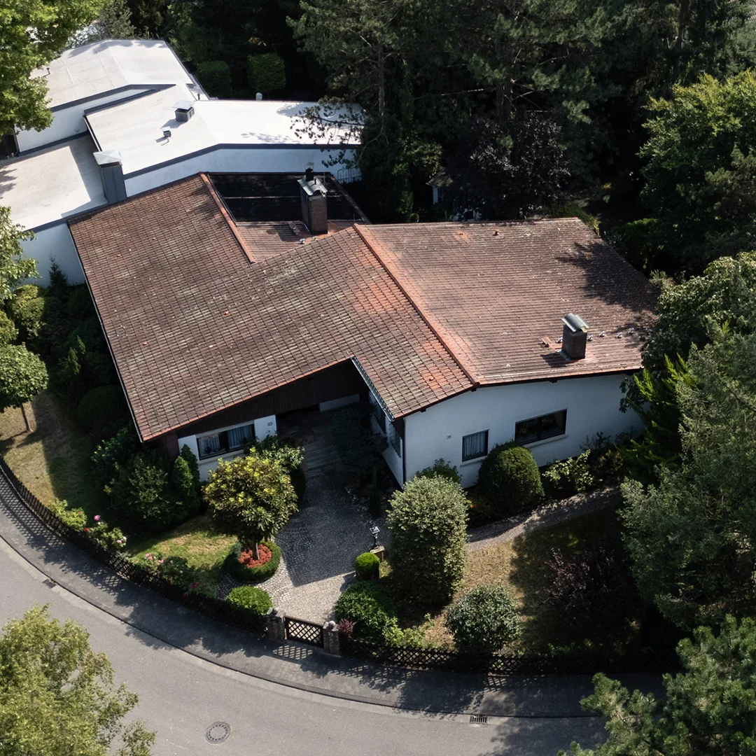 Bungalow mit rotem Satteldach und weißer Fassade, umgeben von Garten mit Büschen und Bäumen an einer Straße.