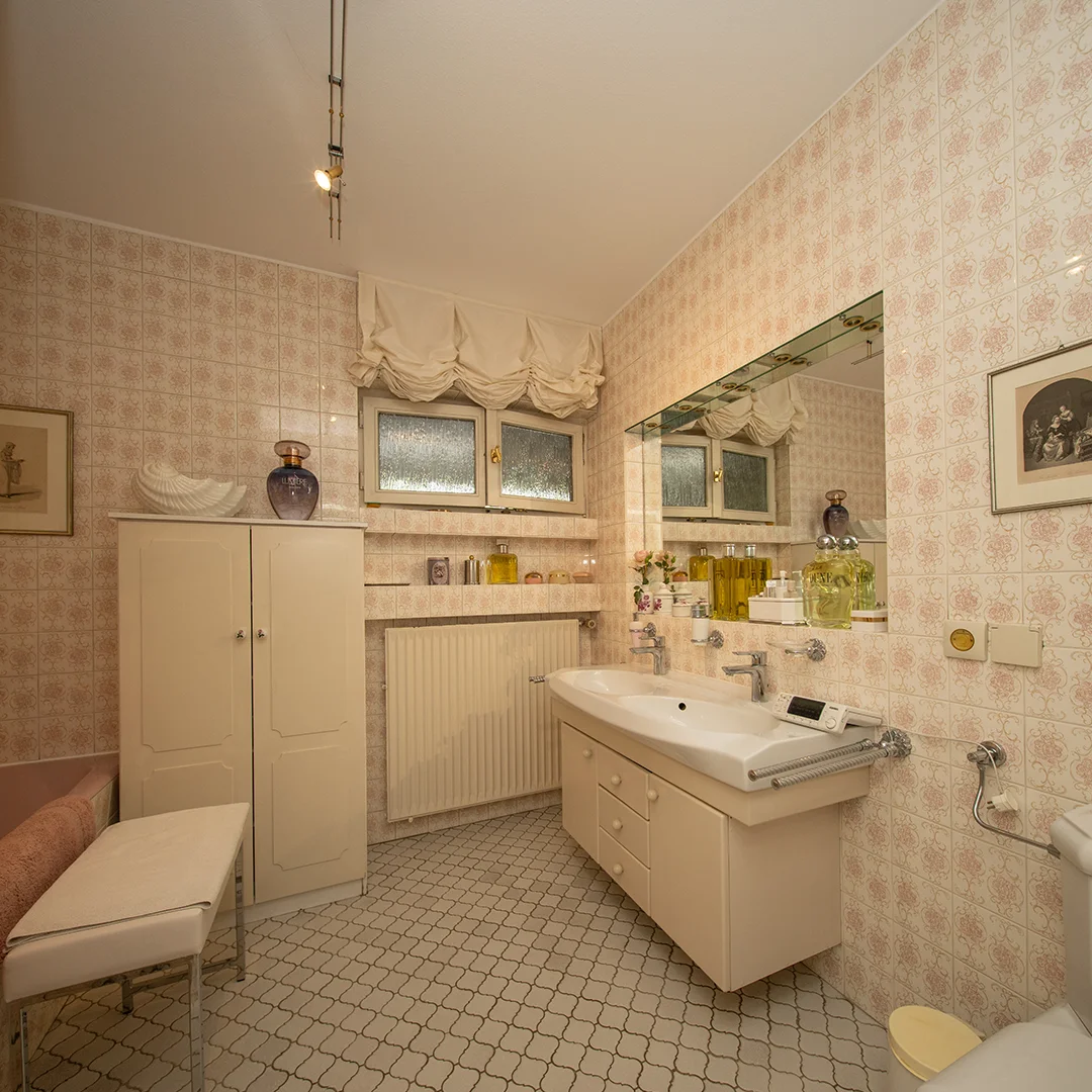 Badezimmer mit großem Spiegel, Waschbecken, Schrank, Fenster mit Gardine und gemusterten Fliesen an Wand und Boden.