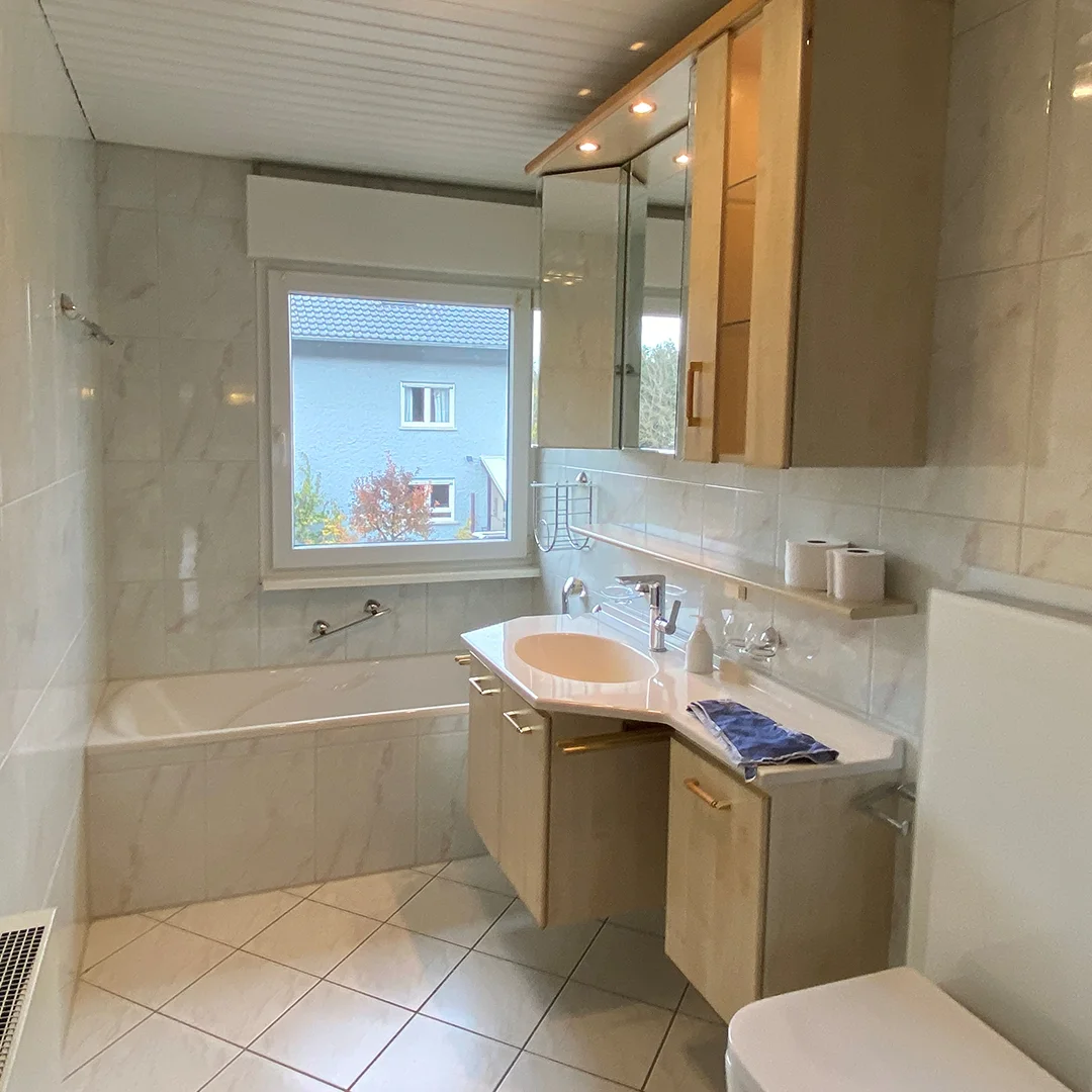 Einfamilienhaus Appenheim Ingelheimer Straße Badezimmer Badezimmer mit Badewanne unter Fenster, Waschbecken mit Spiegel und Holzschränken, Toilette und Fliesenboden.