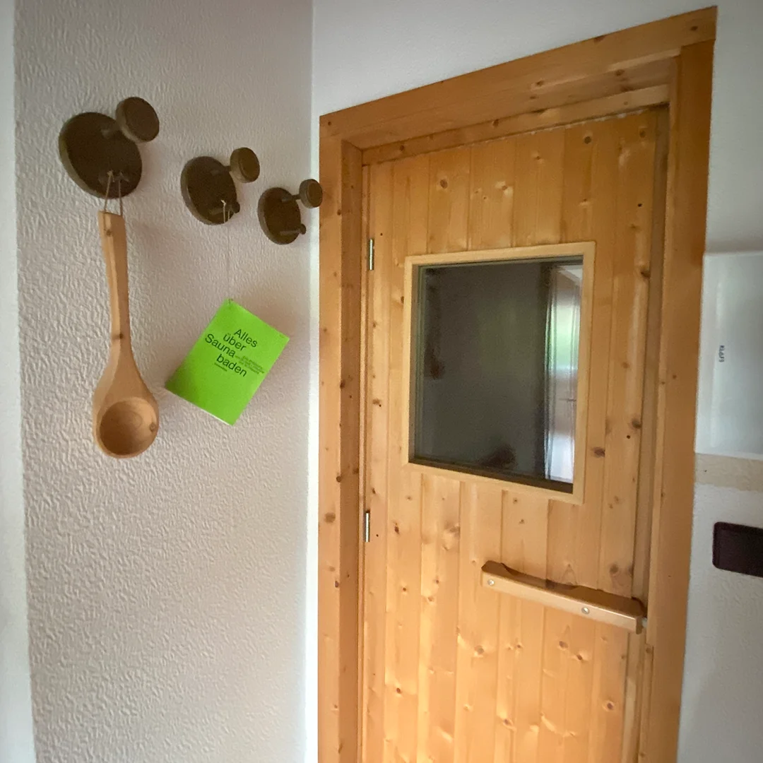 Einfamilienhaus Appenheim Ingelheimer Straße Sauna Holztür mit Fenster in einer Sauna, daneben drei runde Haken an der Wand mit einem Holzlöffel und einem grünen Schild mit Text.