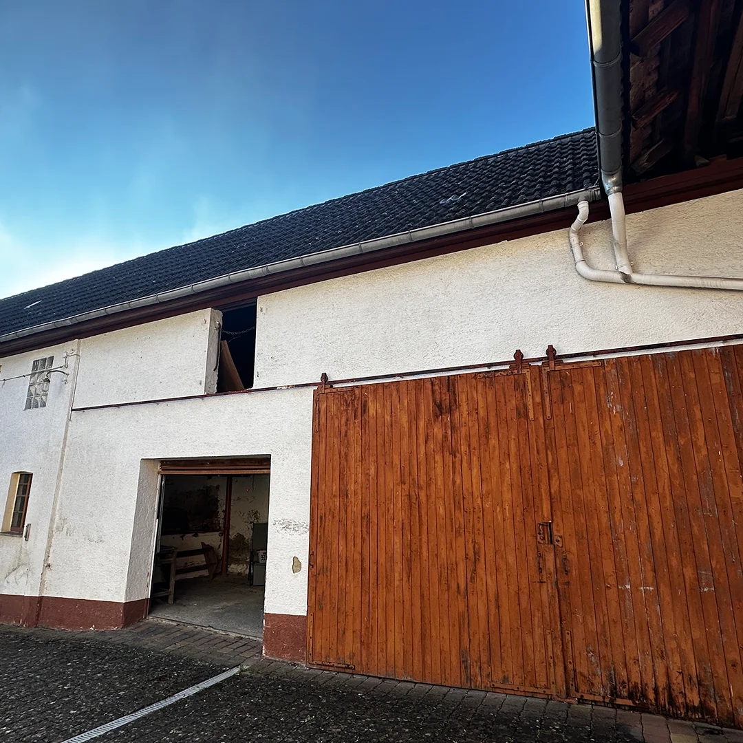 Einfamilienhaus Essenheim Hauptstraße Innenhof Gebäudeabschnitt mit hell verputzter Wandfläche, großem Holztor, offener Durchfahrt, geneigtem Dach, Regenrinne und gepflastertem Boden.