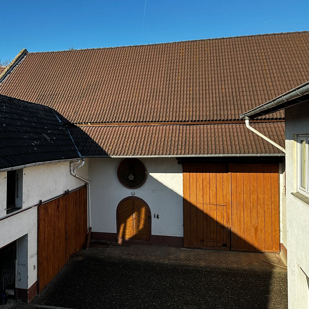 Einfamilienhaus Essenheim Hauptstraße Innenhof 3 Gebäudeabschnitt mit geneigtem Ziegeldach, hell verputzter Wandfläche, großem Holztor, kleiner Holztür, rundem Wandfenster, Regenrinne und gepflastertem Boden.