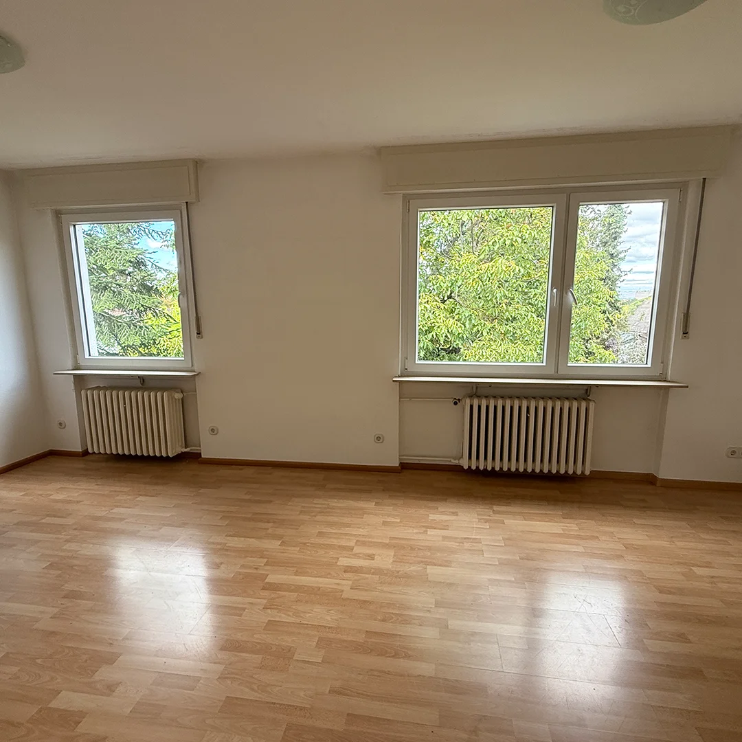 Leerer Wohnraum mit zwei Fenstern und Heizkörpern, heller Holzboden und weißen Wänden.