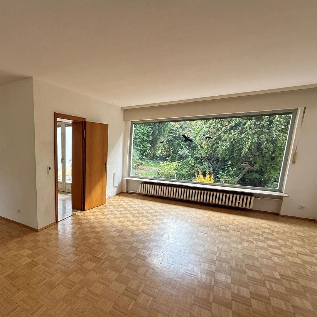 Großes, leeres Wohnzimmer mit Parkettboden, breitem Fenster mit Blick auf Garten und offenem Holztürrahmen zu angrenzendem Raum.