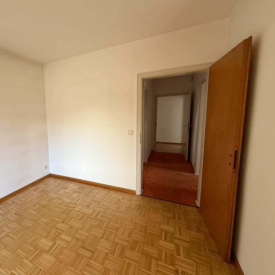 Leerer Raum mit Parkettboden, weißen Wänden und offenem Holztürblatt, Blick in einen Flur mit Teppichboden.