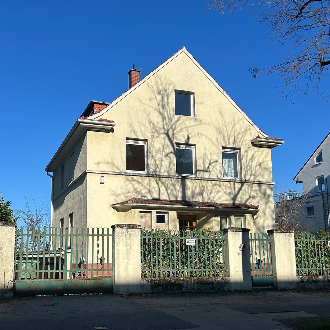 Einfamilienhaus Mainz Wallstraße Außenansicht Gebäude mit hell verputzter Fassade, geneigtem Dach, mehreren rechteckigen Fenstern, Balkonüberdachung, Zaunanlage im Vordergrund und gleichmäßiger Tageslichtbeleuchtung.