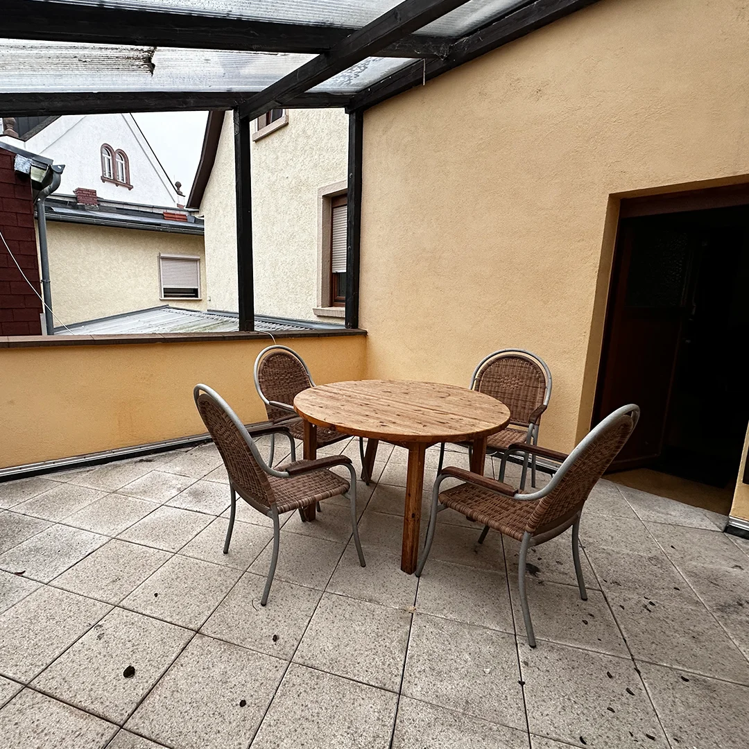 Mehrfamilienhaus Ingelheim Bahnhofstraße Außenbereich Überdachter Bereich mit rundem Holztisch, vier Stühlen, gefliestem Boden und angrenzenden Wandflächen.