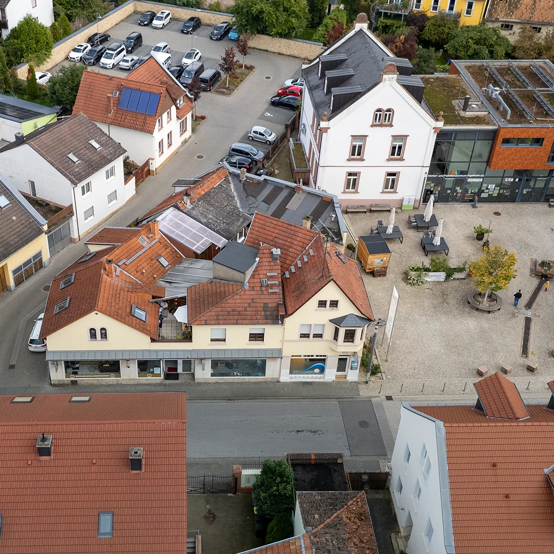 Mehrfamilienhaus Ingelheim Bahnhofstraße Drohnenansicht 2 Luftaufnahme eines Gebäudeensembles mit roten Ziegeldächern, angrenzender Straße, Innenhof, parkenden Fahrzeugen und gepflastertem Platz mit Sitzgelegenheiten.