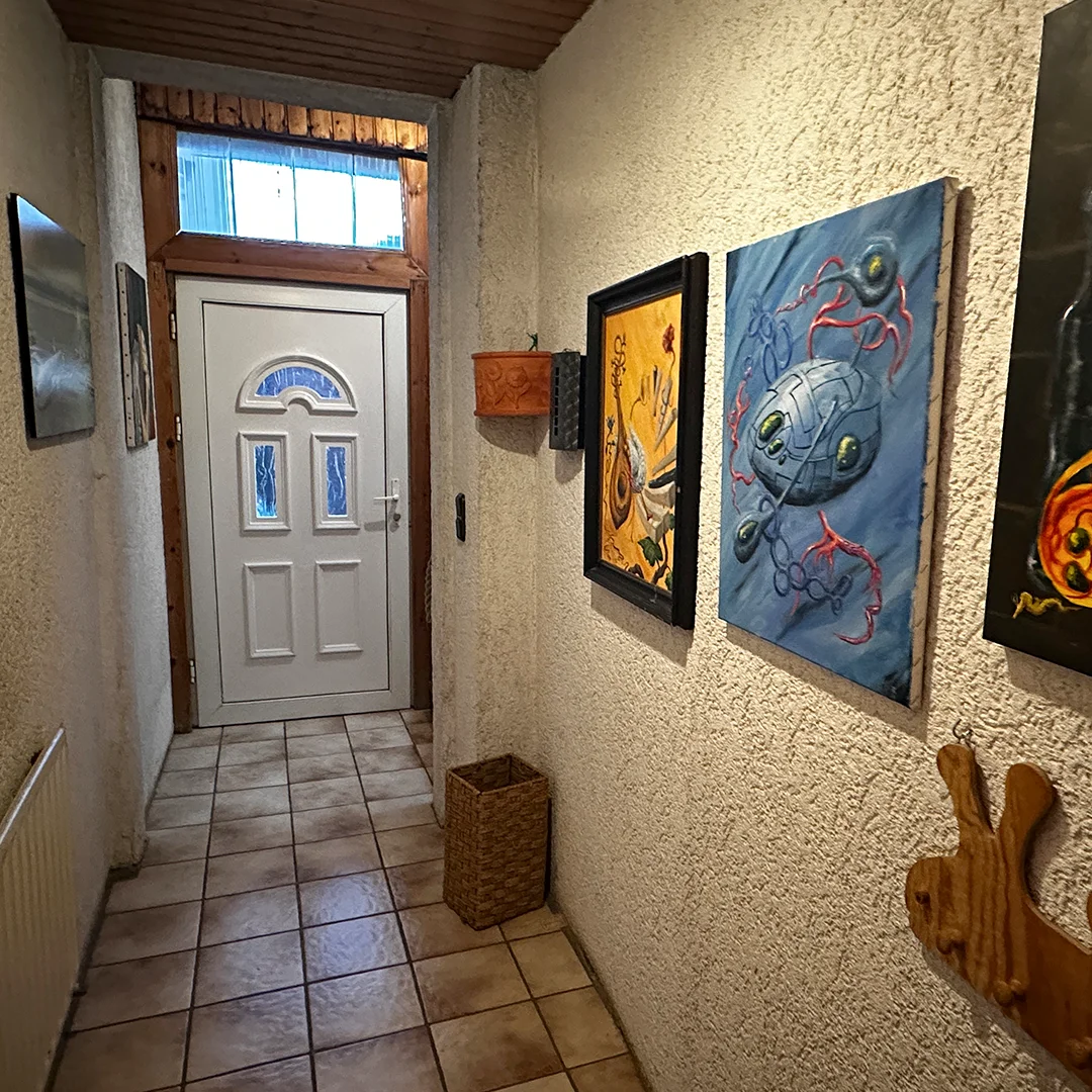 Mehrfamilienhaus Ingelheim Bahnhofstraße Eingangsbereich Schmaler Raum mit gefliester Bodenfläche, strukturieren Wandoberflächen, weißer Tür mit Glaseinsatz, Holzeinfassung, Wandbildern und Deckenverkleidung aus Holz.