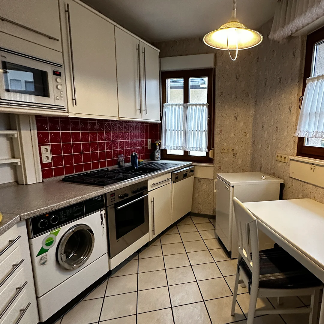 Mehrfamilienhaus Ingelheim Bahnhofstraße Küche Schmaler Raum mit gefliestem Boden, Einbauküche mit hellen Schränken, roten Wandfliesen, Arbeitsfläche mit Kochfeld, Haushaltsgeräten, Fenster mit Gardinen, Deckenleuchte sowie Tisch und Stuhl.