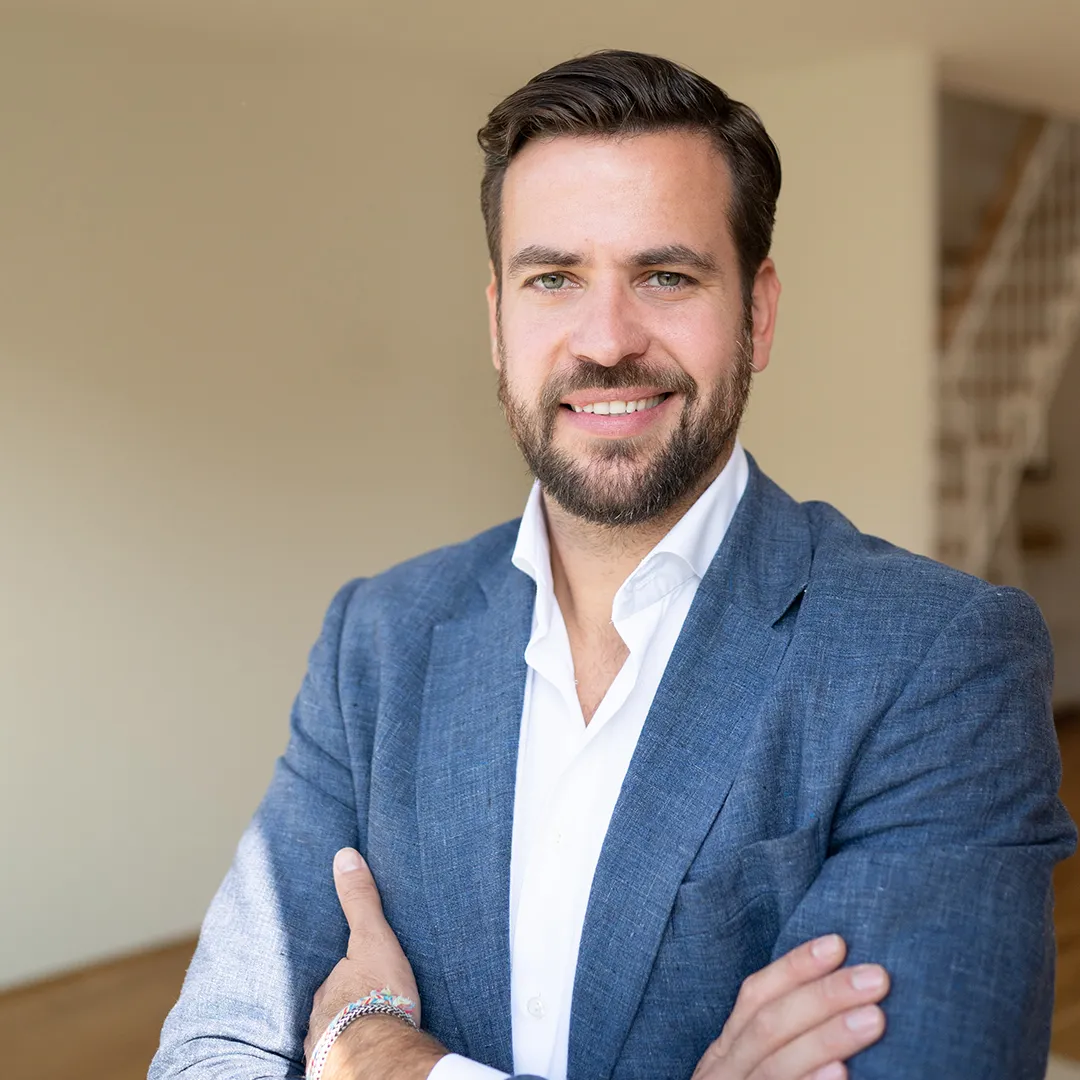 Mann mit blauem Sakko und weißem Hemd steht mit verschränkten Armen vor einer hellen Wand und Treppe.