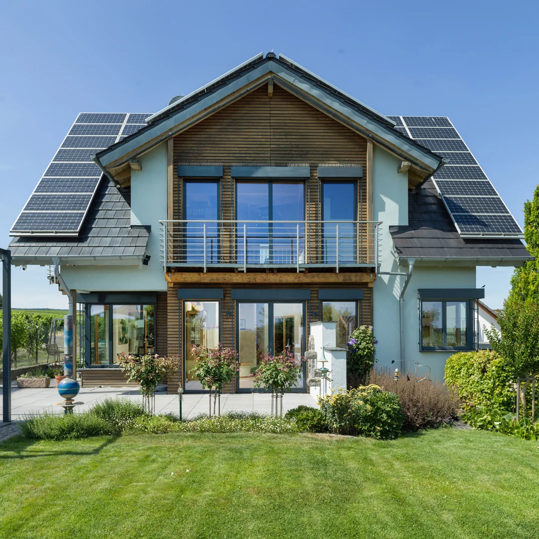 Gebäude mit Satteldach, Holzfassade, Balkon, großen Fensterflächen und Solarmodulen auf dem Dach, davor Grünfläche.