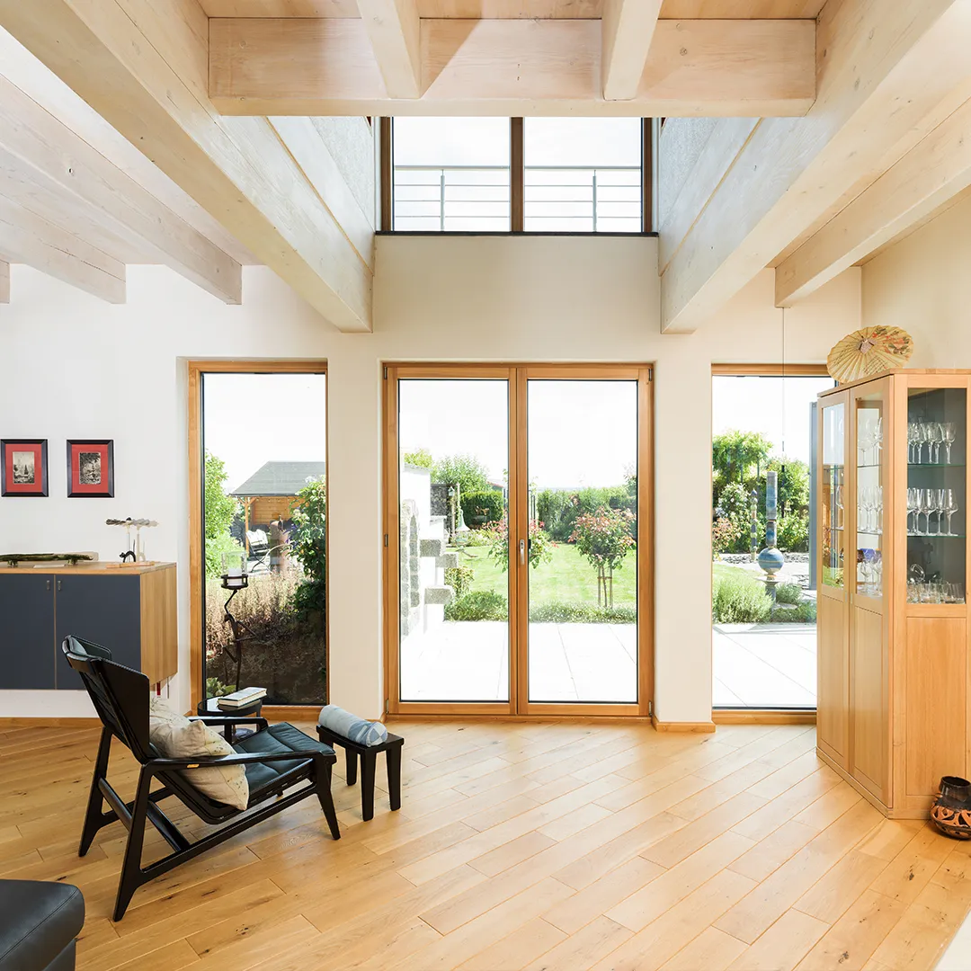 Raum mit hellem Holzboden, sichtbaren Deckenbalken und großflächigen Glas- und Fensteröffnungen.