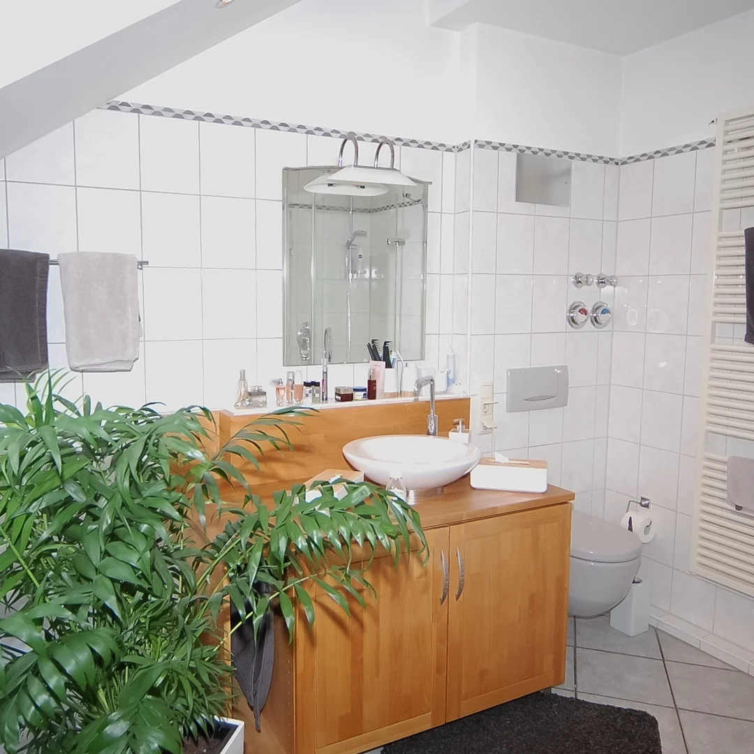 Badezimmer mit Holzwaschtisch, rundem Aufsatzwaschbecken, Spiegel, weißen Fliesen und großer grüner Pflanze im Vordergrund.