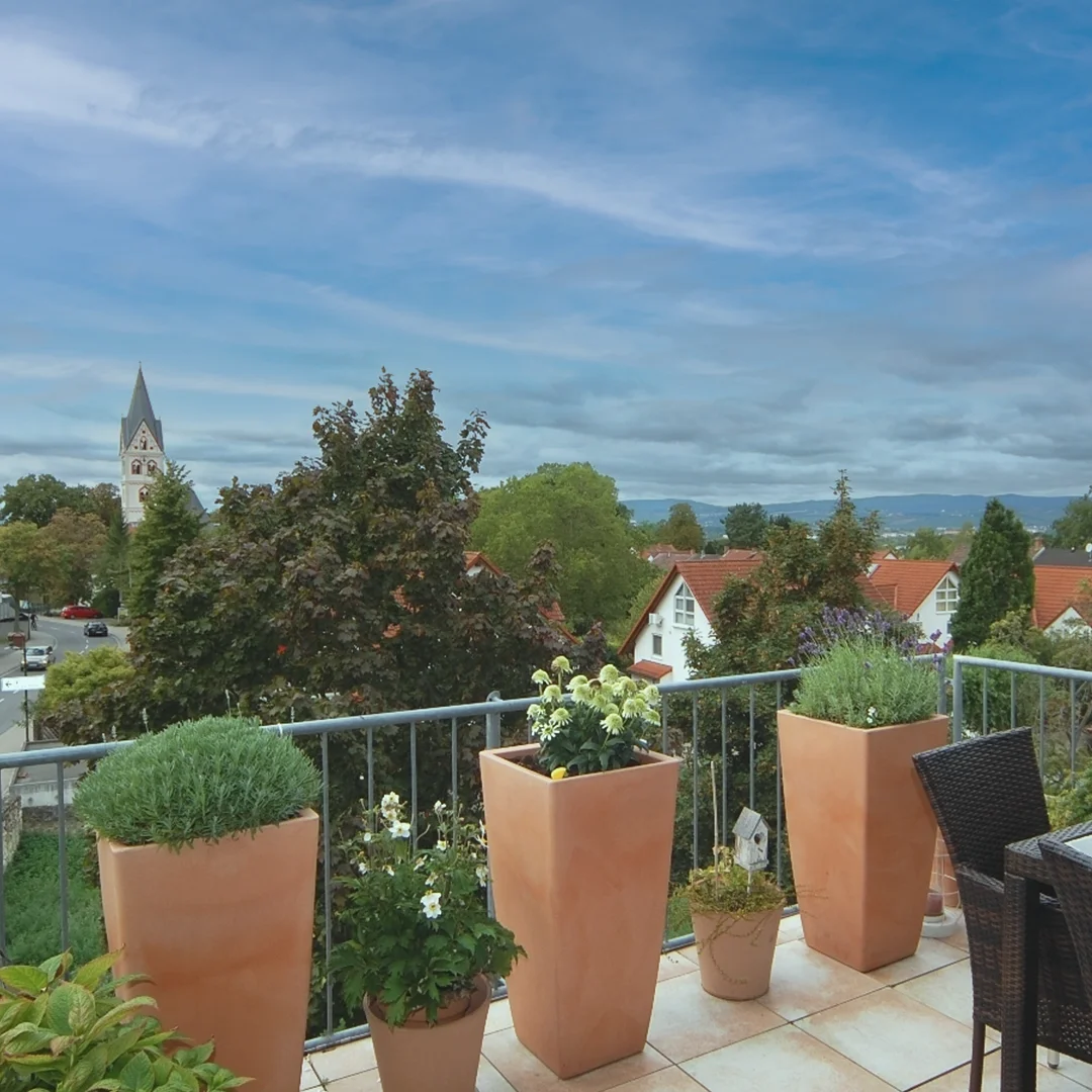 Balkon mit mehreren großen und kleinen Pflanzkübeln, im Hintergrund Bäume, Häuser und ein Kirchturm unter bewölktem Himmel.