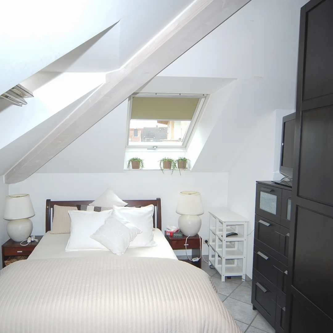 Schlafzimmer mit Dachschräge, Doppelbett mit mehreren Kissen, zwei Nachttischen mit Lampen, Fenster mit drei kleinen Pflanzen und schwarzem Schrank mit Fernseher.