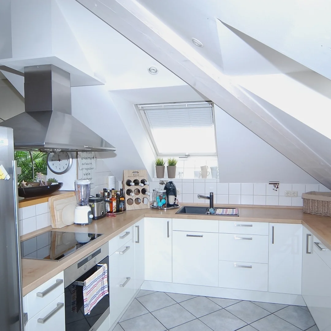 Moderne Dachschrägenküche mit weißen Schränken, Holzarbeitsplatte, Edelstahl-Dunstabzugshaube und eingebautem Backofen.