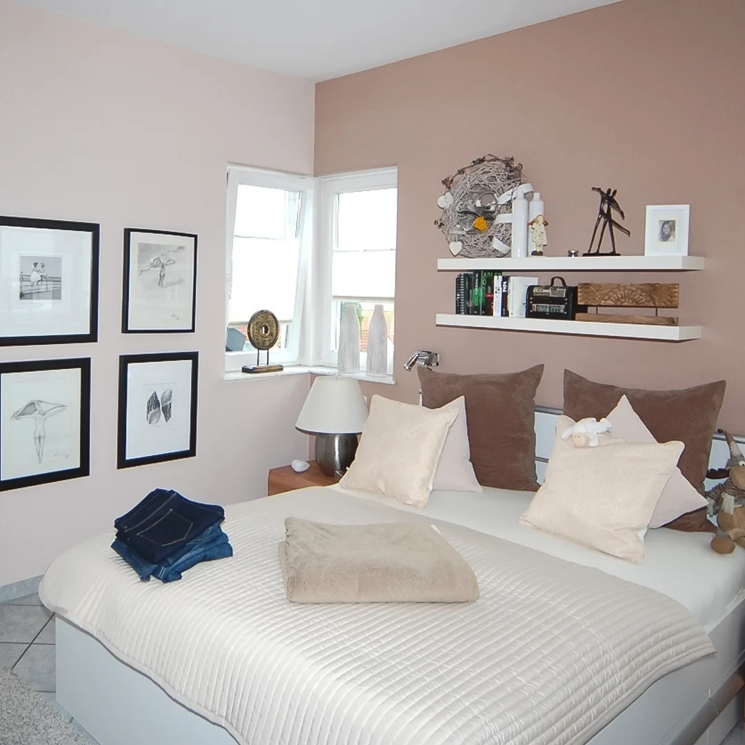 Schlafzimmer mit Doppelbett, mehreren Kissen, zwei Regalbrettern mit Dekoration und Bildern an der Wand neben einem Fenster.