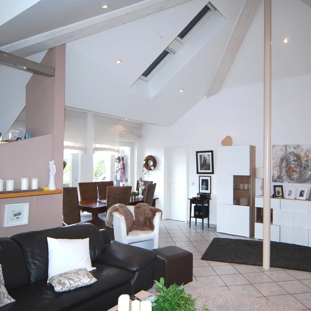 Offenes Wohnzimmer mit schwarzem Ledersofa, weißem Sessel, Esstisch mit Stühlen und moderner weißer Wohnwand unter schräger Decke mit Dachfenstern.