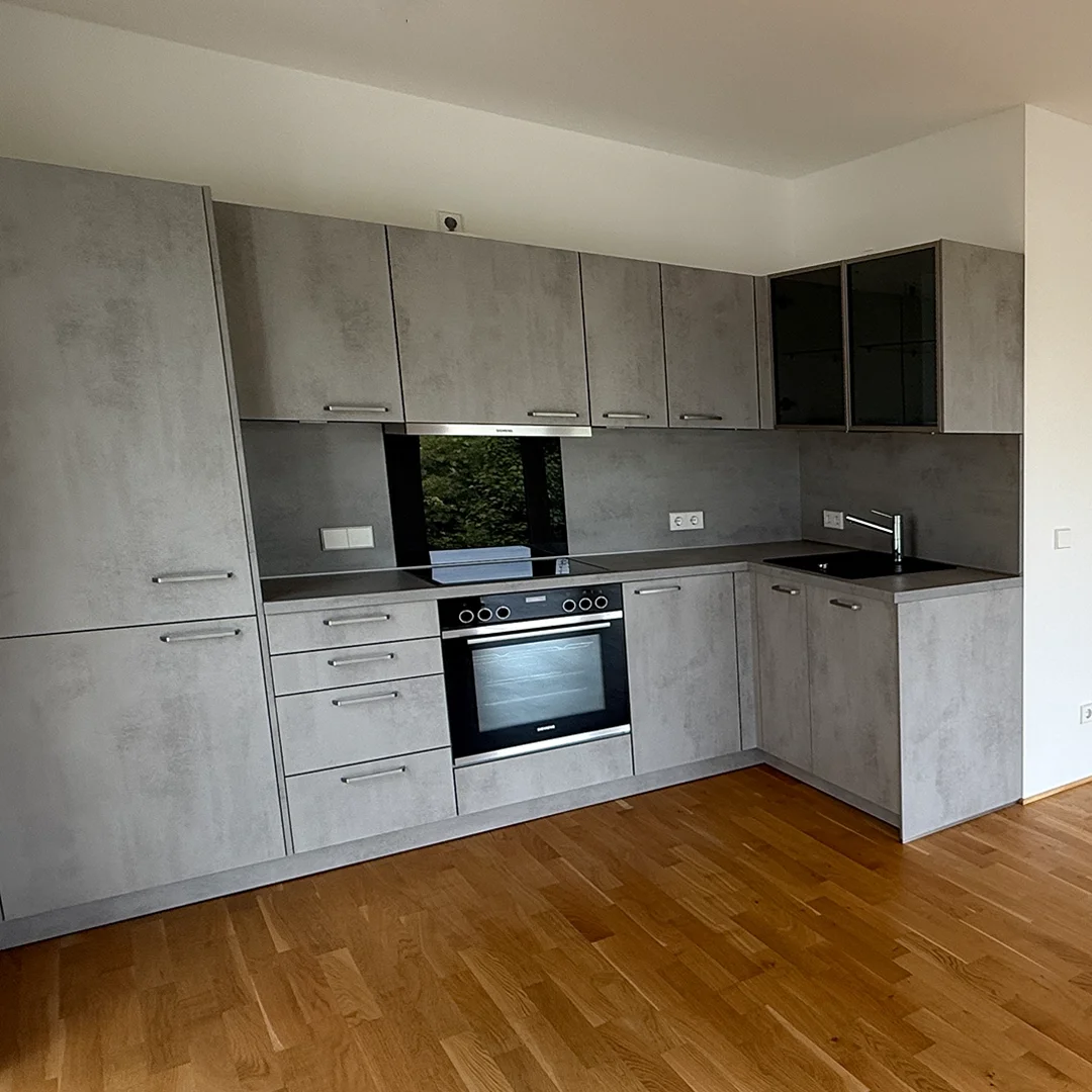 Moderne Küche mit grauen Fronten, eingebautem Backofen, Cerankochfeld und Spüle in L-Form, heller Holzboden.