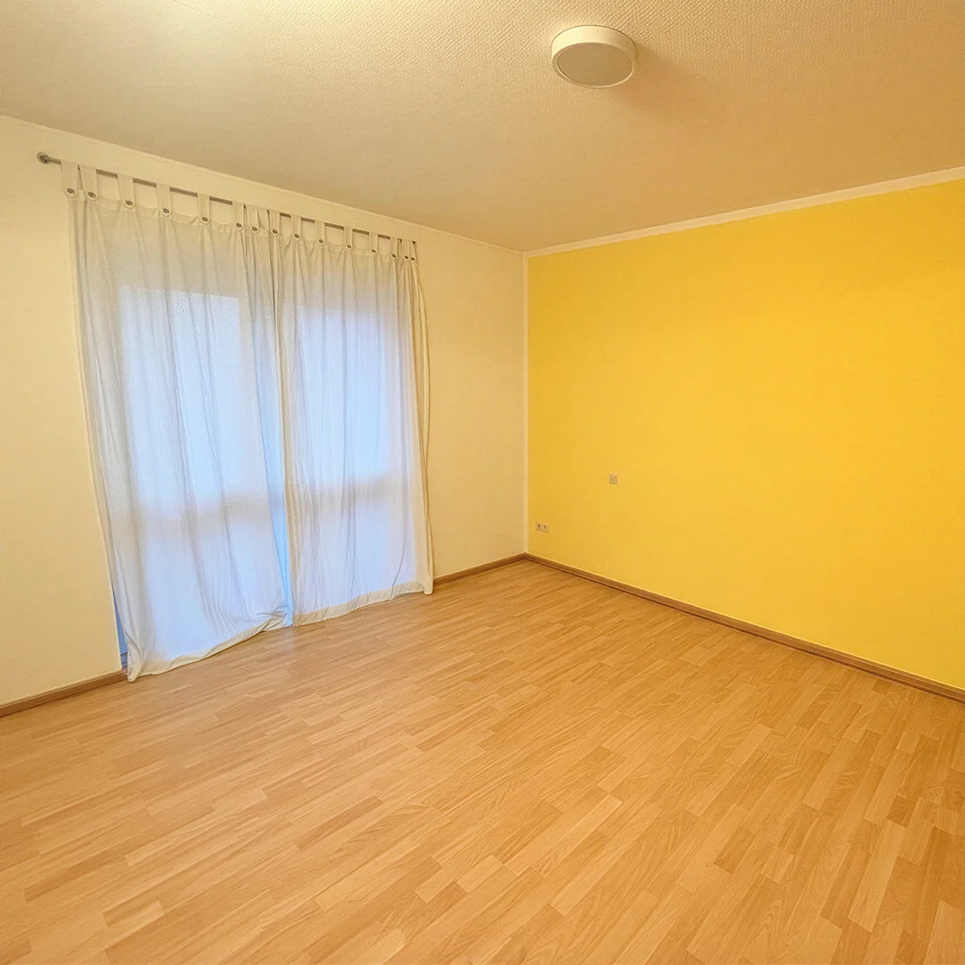 Leerer Raum mit hellem Holzboden, gelber Wand und Fenster mit weißen Vorhängen