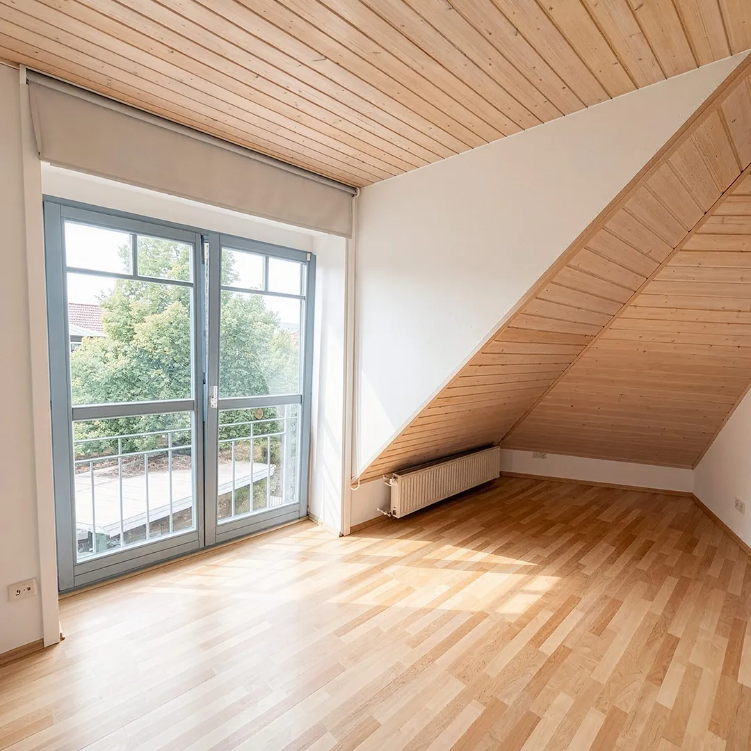 Helles Schlafzimmer mit Parkettboden, holzverkleideter Dachschräge und großer Balkontür mit Blick ins Grüne.