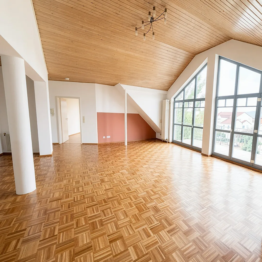 Großzügiges, helles Wohnzimmer mit Parkettboden, holzverkleideter Dachschräge und großer Fensterfront mit Zugang zum Balkon.